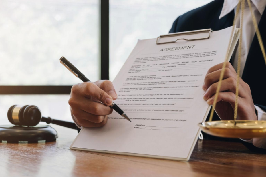 Lawyer signing a legal agreement document beside a judge’s gavel and scales of justice.
